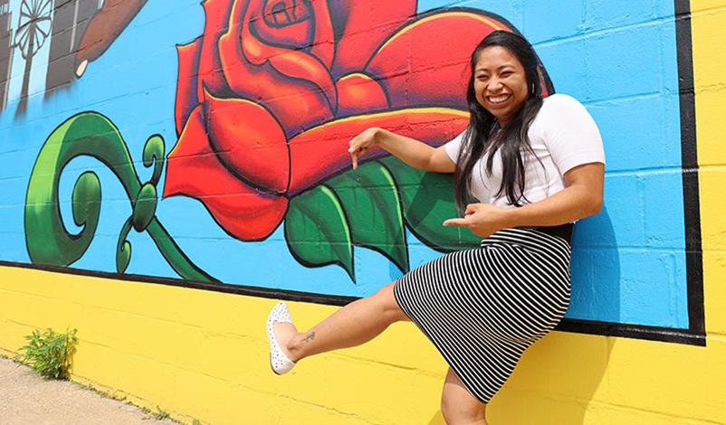 Alma standing in front of a Cedar Rapids mural, smiling and pointing to her similar ankle tattoo